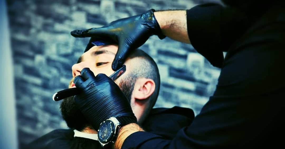 Een barber werkt met een scheermes de baard bij; zwarte handschoenen en het knipwerk van dichtbij.